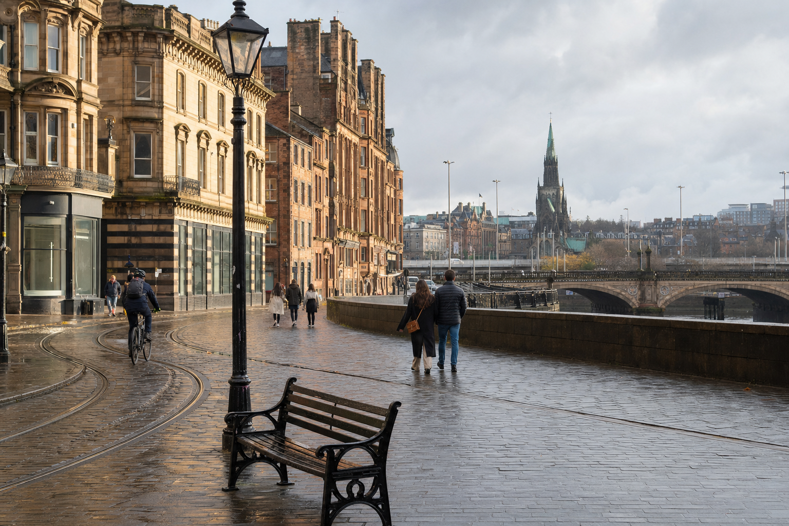 People walk and cycle along a wet cobblestone street in a historic city with a church spire in the distance.