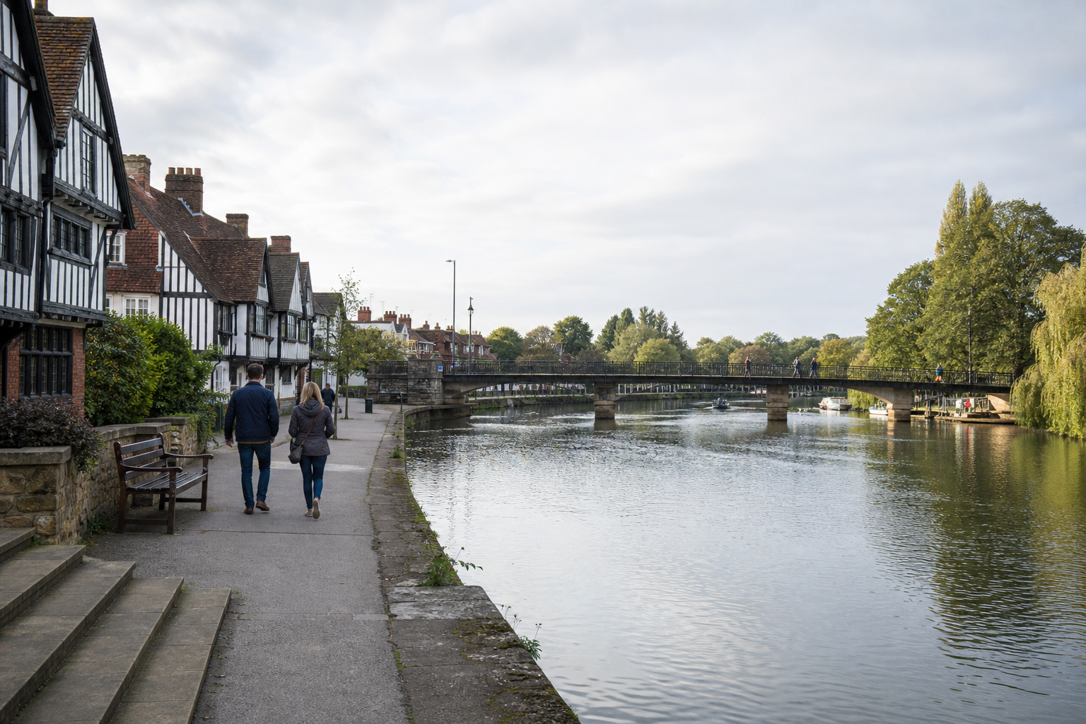 A couple walks along a riverside path beside historic timber-framed buildings, with a bridge crossing the river ahead.