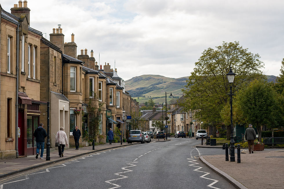 A quiet British village street with stone buildings, a few pedestrians, and hills in the background.