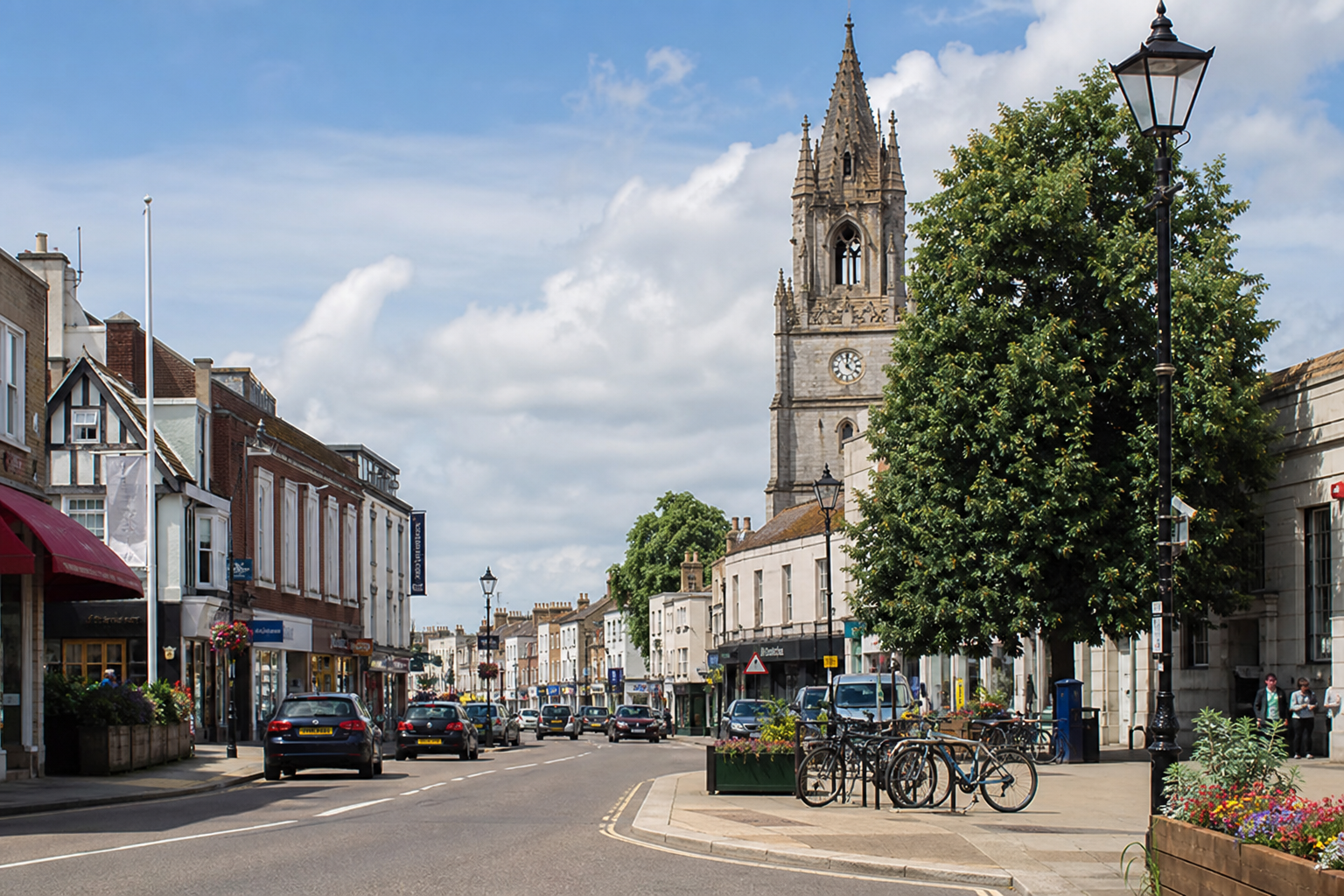 A historic street with a clock tower, shops, parked cars, and a tree on a sunny day.