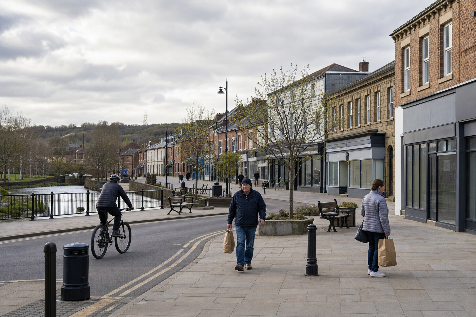 People walking and cycling along a riverside street lined with trees and buildings under a cloudy sky.