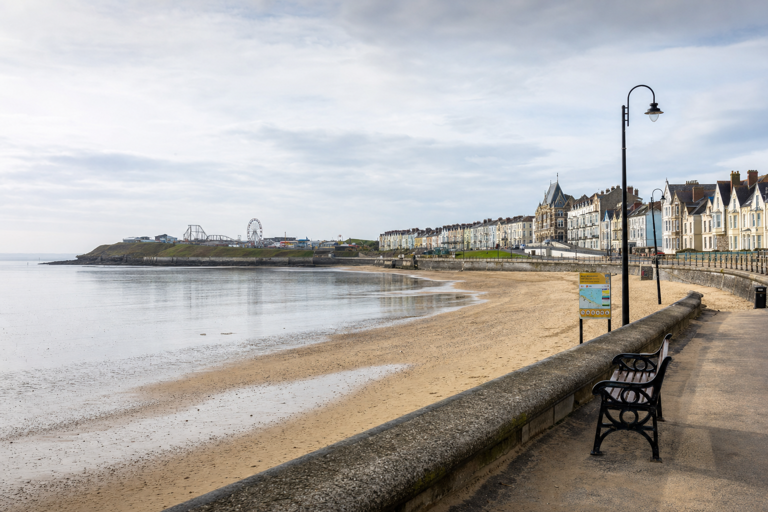 A coastal promenade with Victorian houses, a sandy beach, and an amusement park in the distance under a cloudy sky.