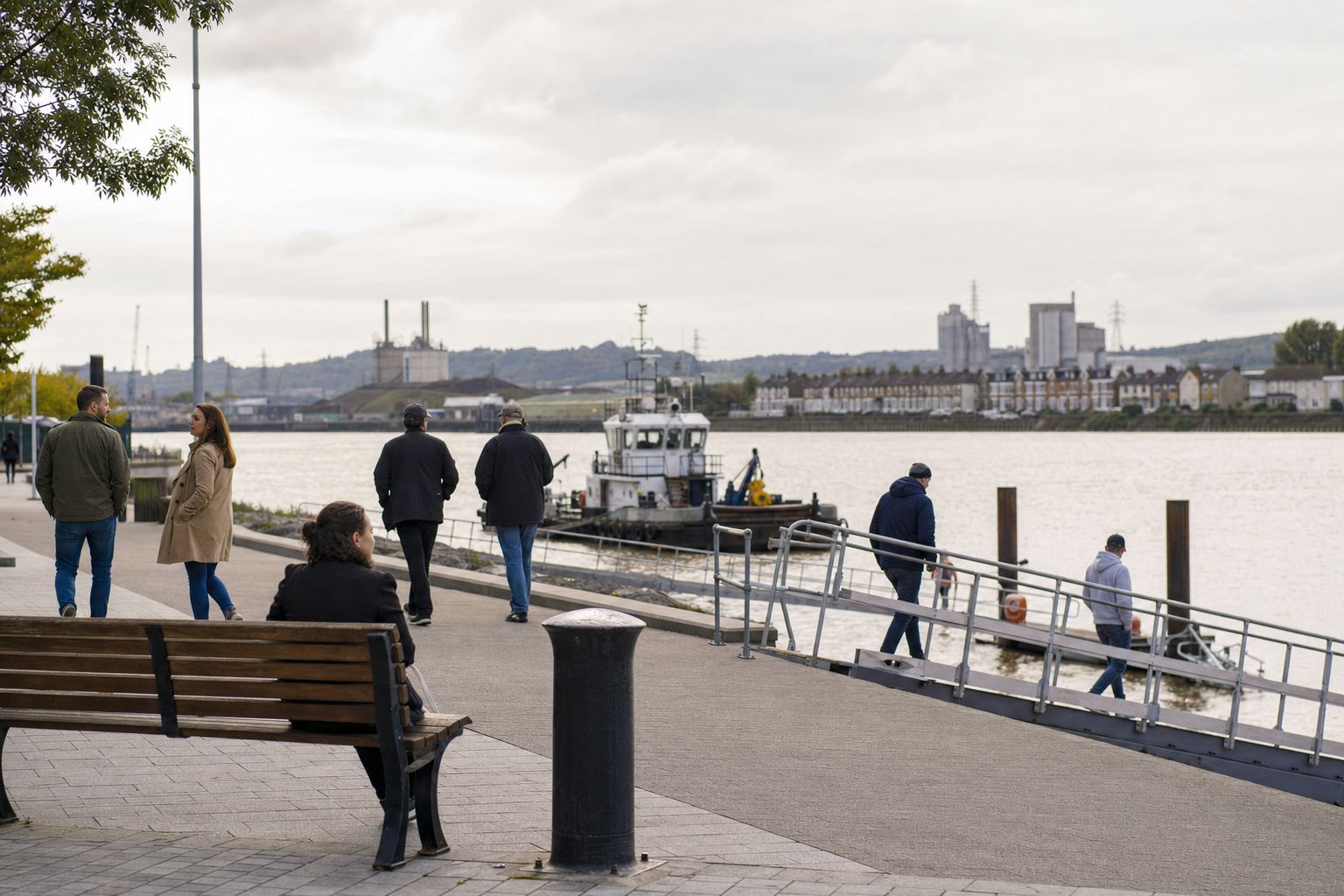 People walking along a riverside path with a boat on the water and industrial buildings in the background.