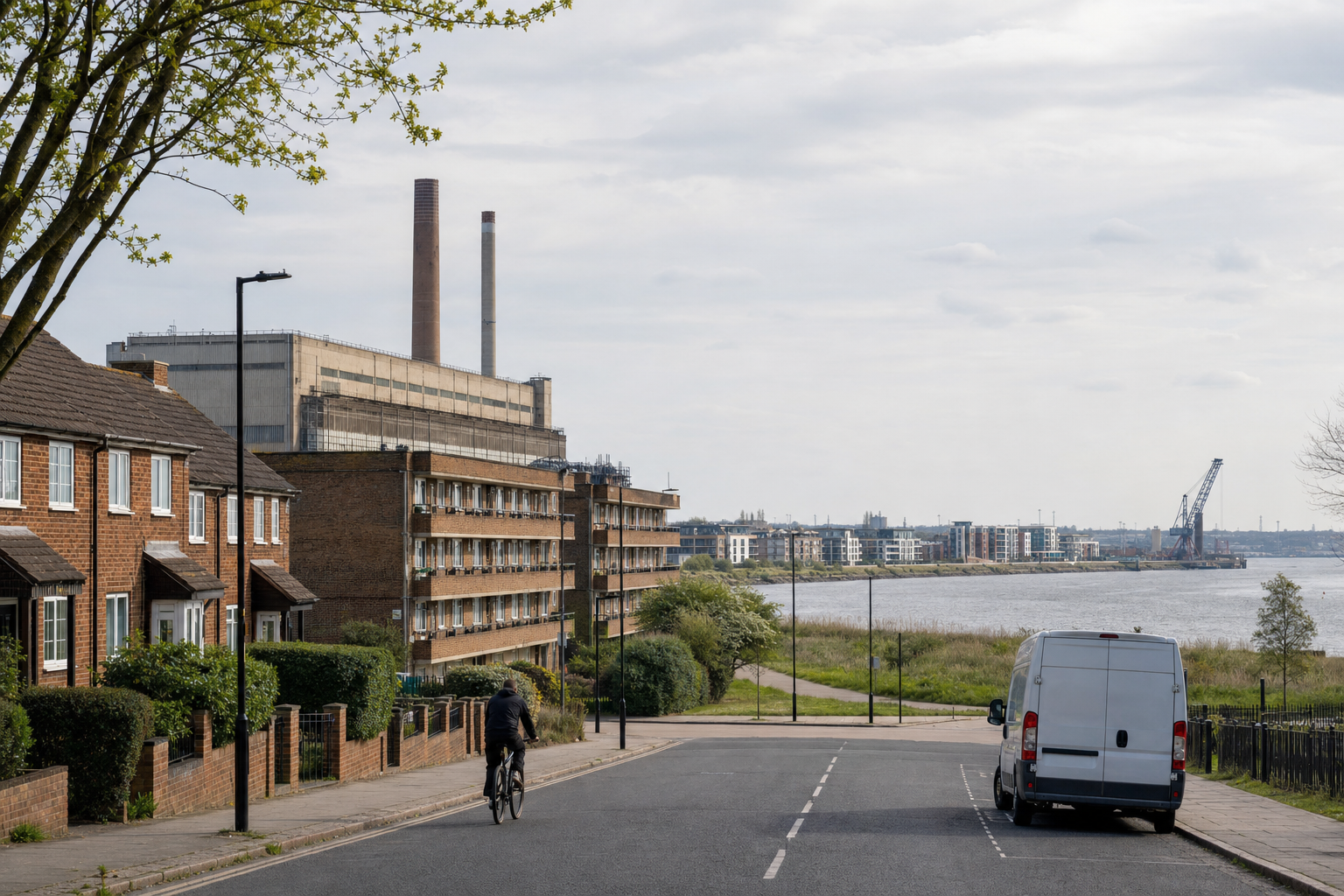 Cyclist riding towards a power station by the river, with terraced houses and a parked van nearby.