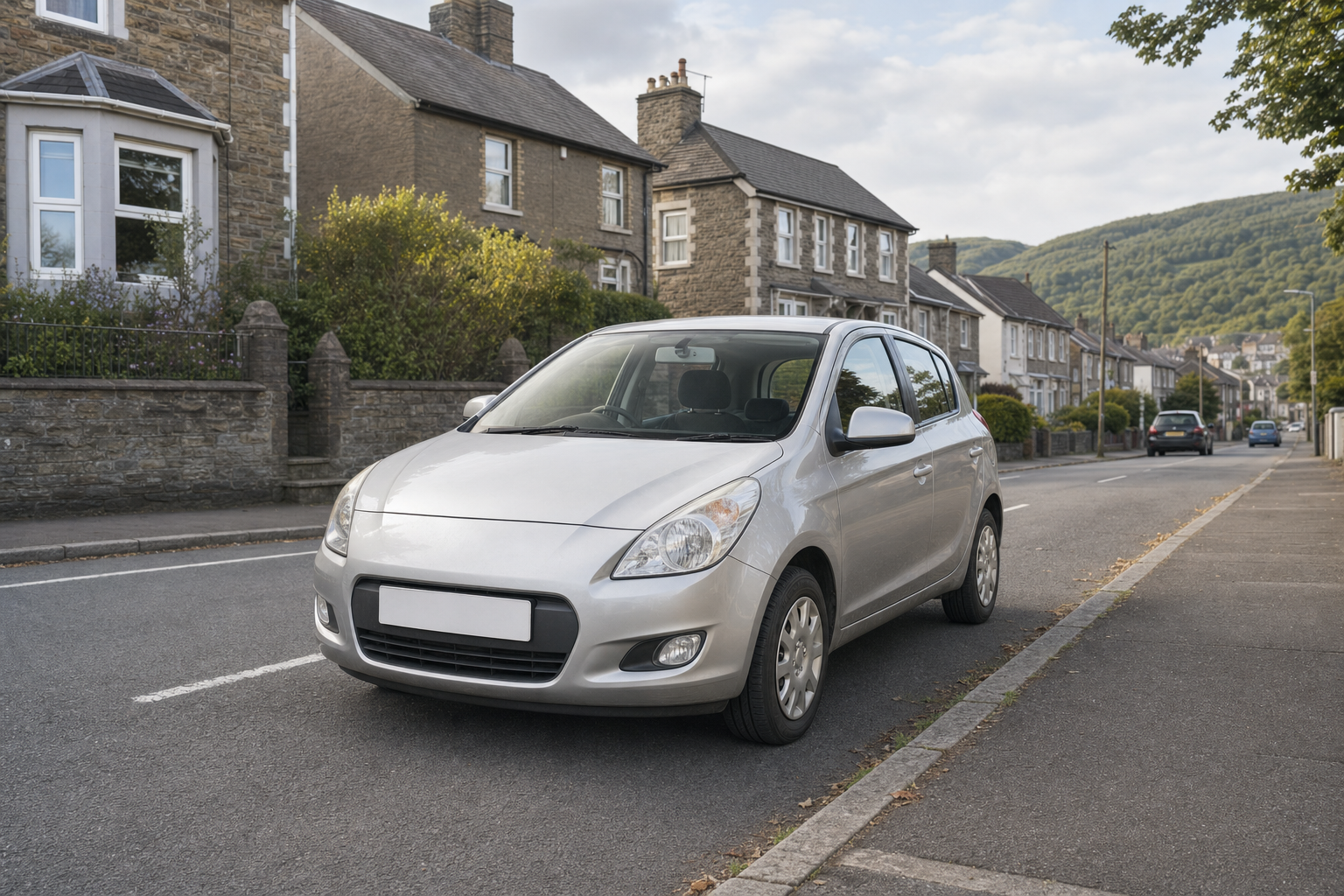 A silver car parked on a suburban street with stone terraced houses and a distant view of green hills.