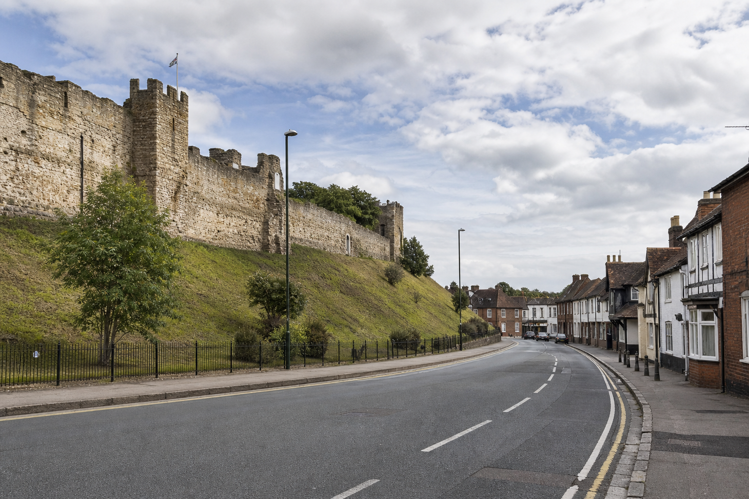 Historic stone castle wall alongside a quiet street lined with traditional houses under a partly cloudy sky.