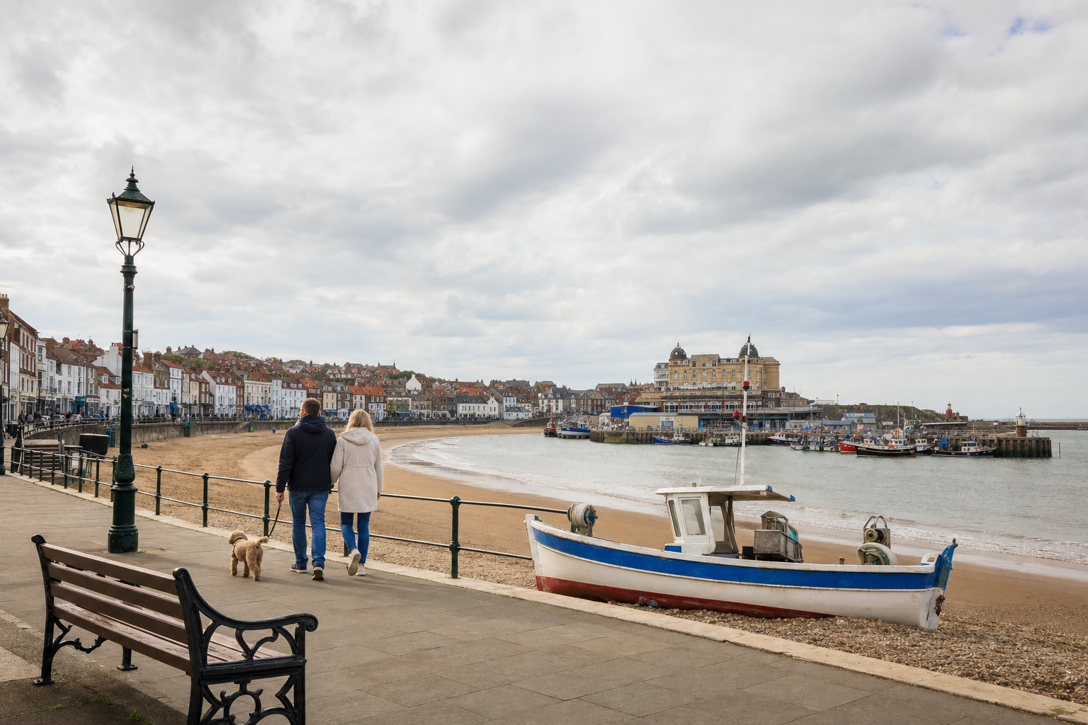 A couple walks a dog along a coastal promenade, with a boat and harbour visible in the background under a cloudy sky.