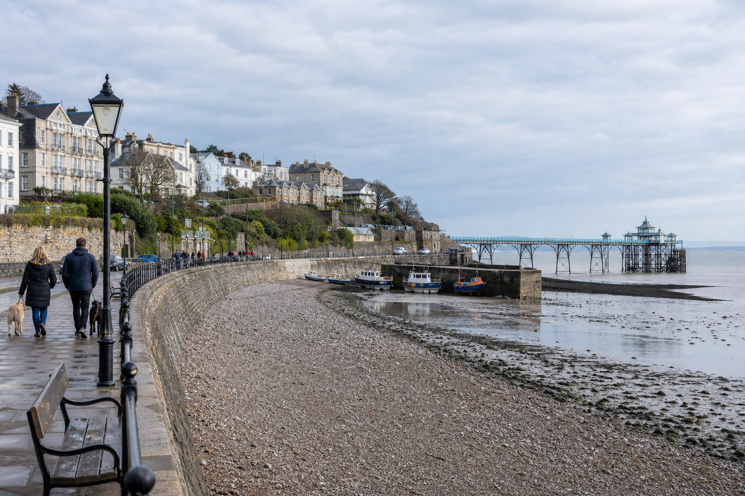 Coastal promenade with people walking dogs beside a pier and buildings in the background.