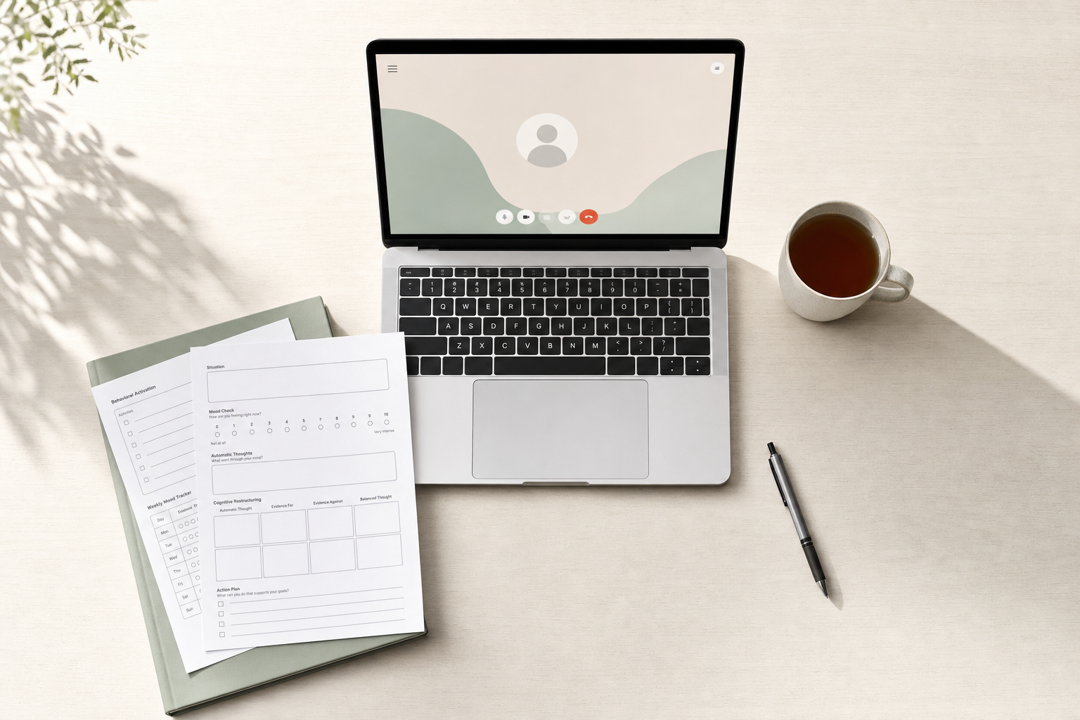 A laptop displaying a video call screen, alongside a notebook, printed worksheets, a cup of tea, and a pen on a light desk.