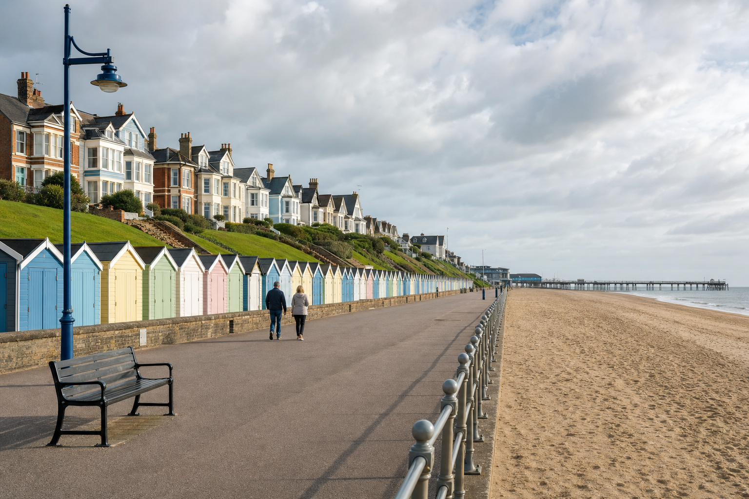 Seaside promenade with colourful beach huts, pedestrians, and classic homes under a cloudy sky.