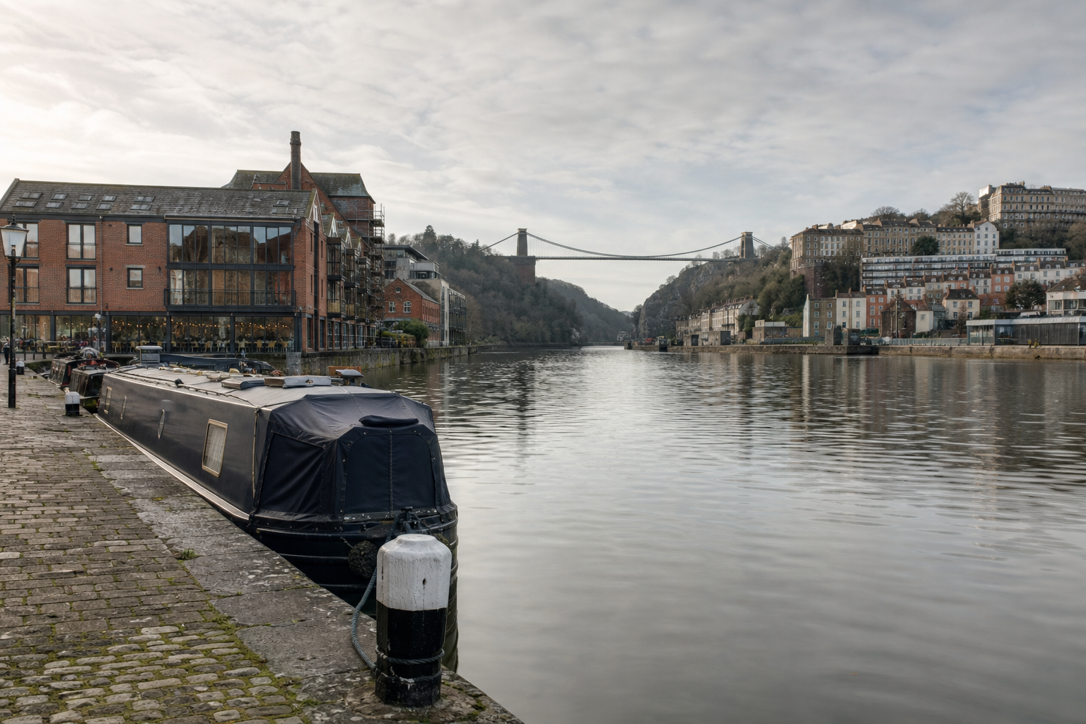 Canal barge moored alongside a cobbled quay with the Clifton Suspension Bridge in the background.