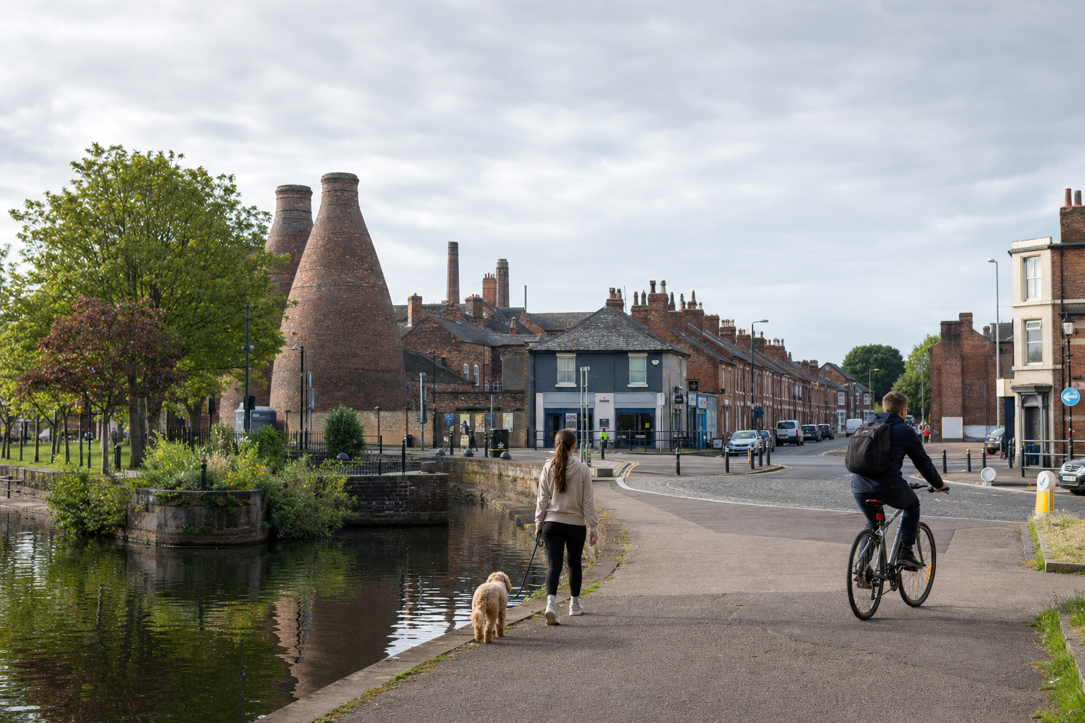 A woman walks her dog beside a canal near brick bottle kilns as a man cycles in an urban area.