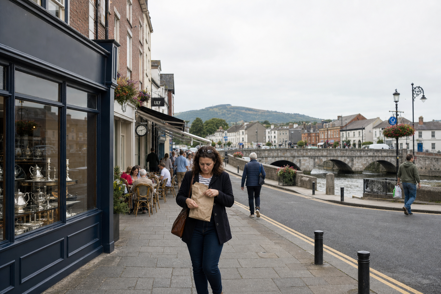 A woman walks down a street holding a brown paper bag, with a display of silverware and a bridge in the background.