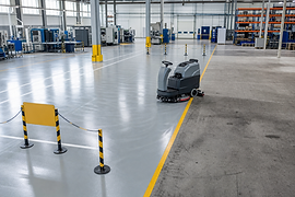 An industrial floor cleaner sits in a large, empty warehouse with marked safety lines.