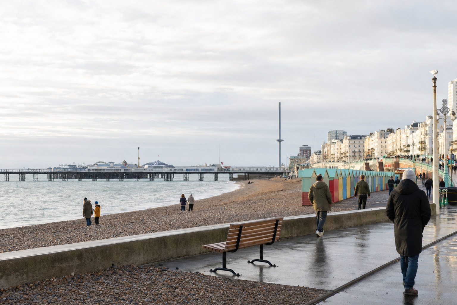 Brighton beach scene with a pier, colourful beach huts, and people walking along the promenade under a cloudy sky.