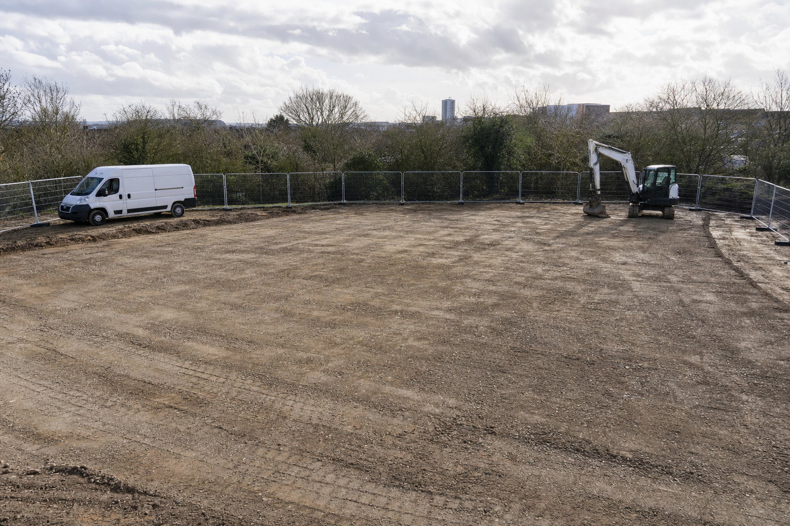 A fenced, levelled construction site with a white van and an excavator under a cloudy sky.