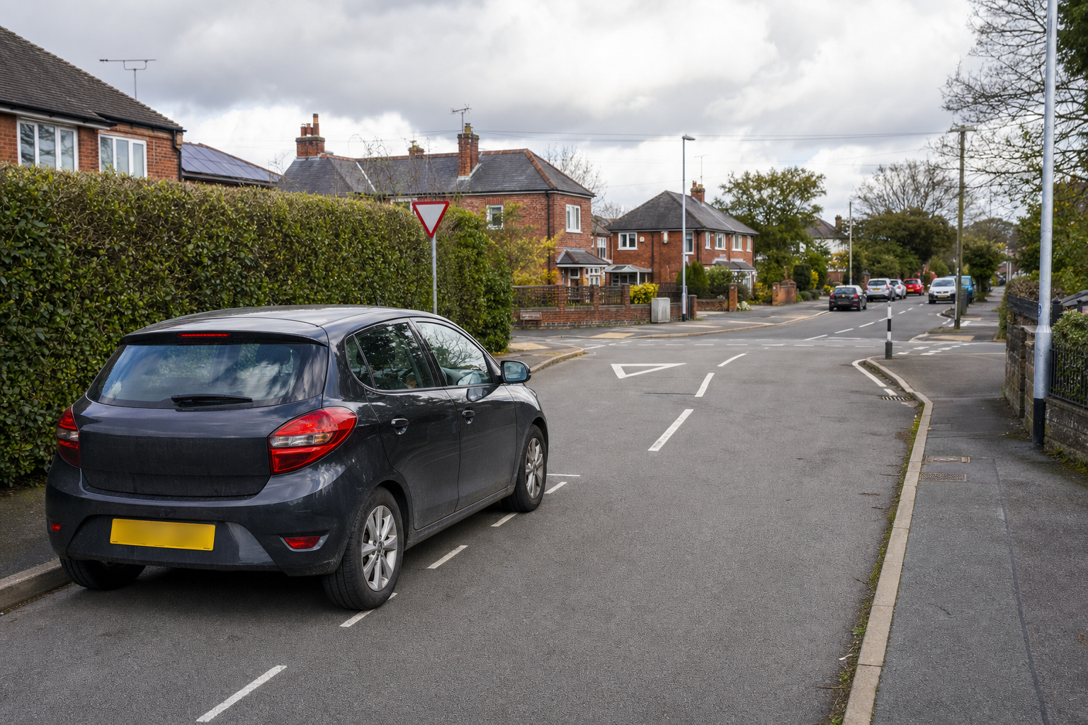 A parked black car on a quiet suburban street with a yield sign and detached houses in the background.