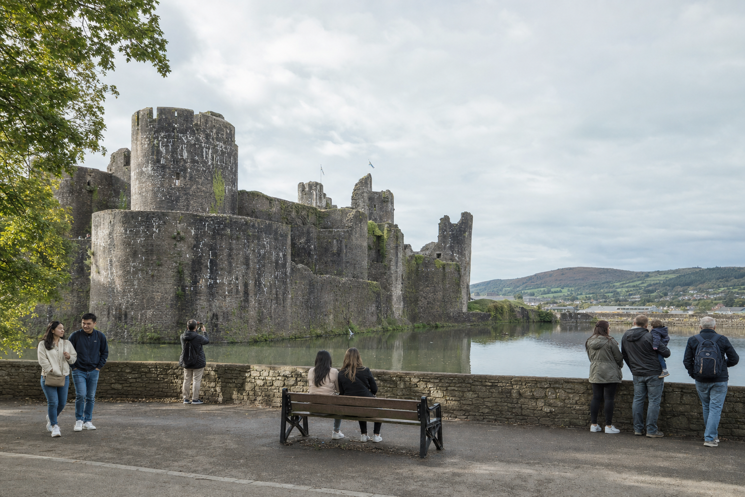 People enjoying the view of a historic castle by a river, with hills and cloudy sky in the background.