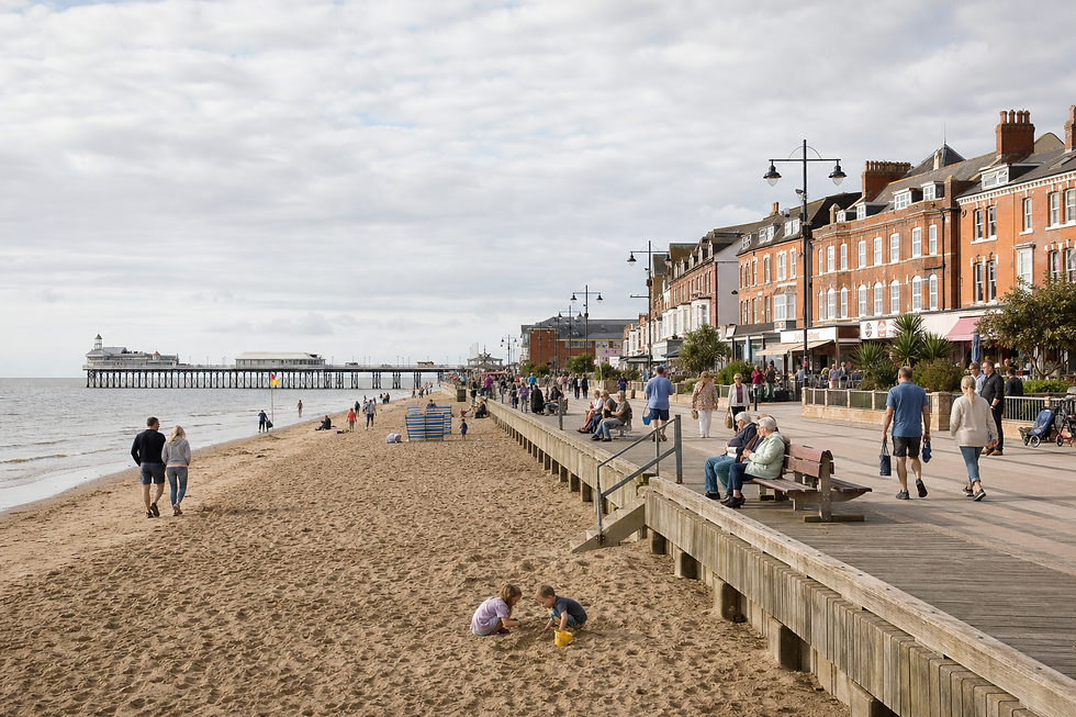 People enjoying a sunny day on the beach and promenade near a pier, with shops and cafes lining the street.