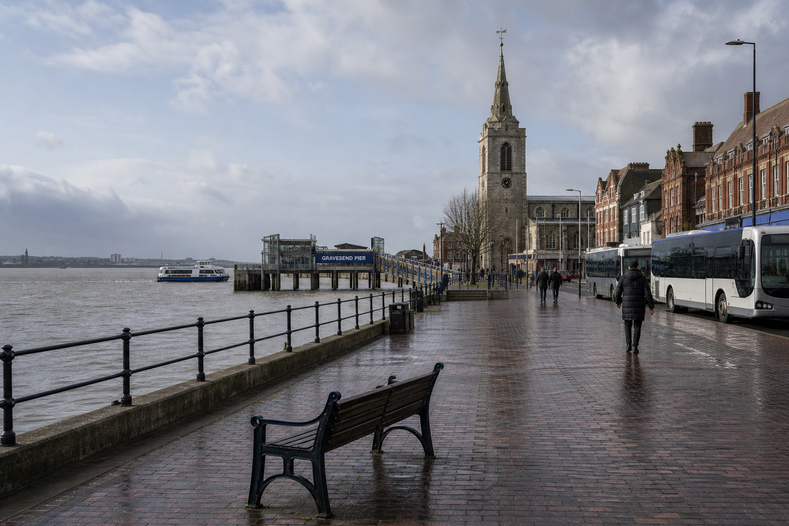 A riverside view with Gravesend Pier, a historic church, and buses lined along a wet promenade under a cloudy sky.