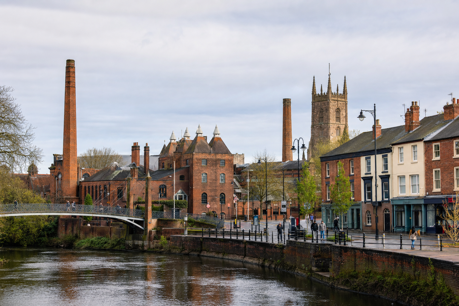 Historic buildings and a church tower beside a river in a quaint British town, with a pedestrian bridge in the foreground.