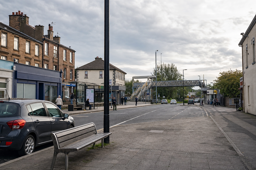 A quiet street scene with a parked car, bench, bus stop, and overhead footbridge under a cloudy sky.