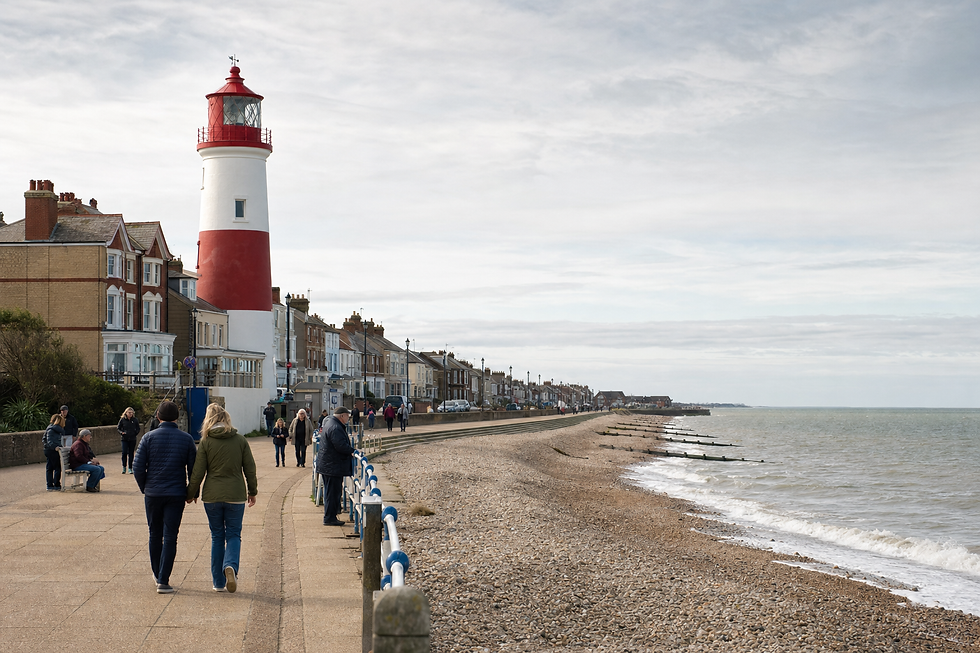 People walking along a seaside promenade with a red and white lighthouse and pebble beach in the background.