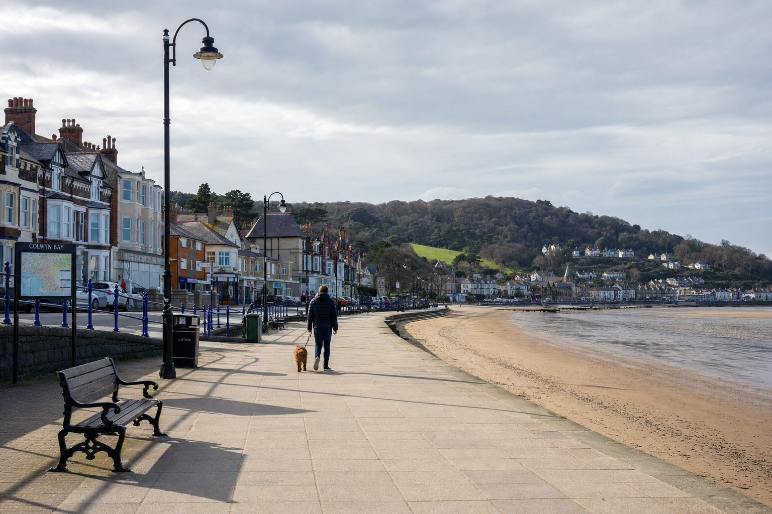 A person walks a dog along a seafront promenade in Colwyn Bay with hillside houses in the background.