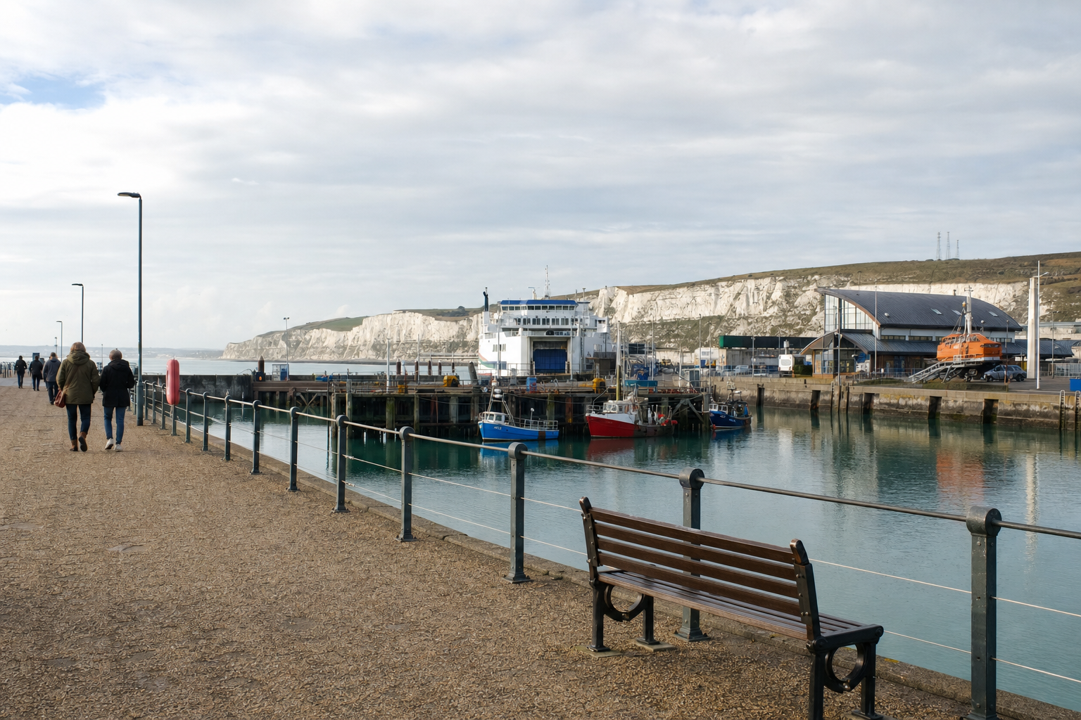 A coastal harbour scene with people strolling along the promenade and boats docked by the water.