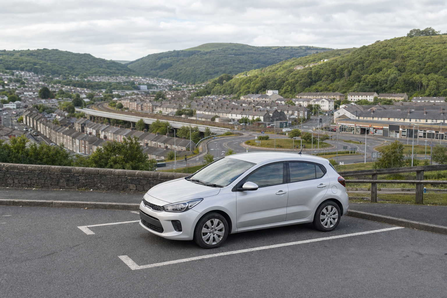 A silver hatchback car parked in a hilltop space overlooking a valley townscape with green hills in the background.