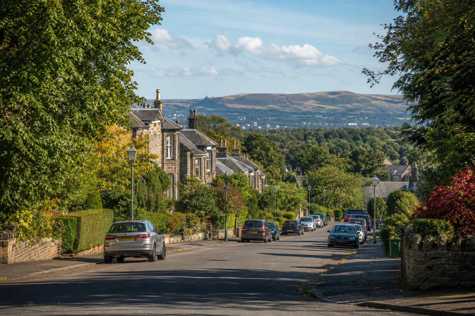 Tree-lined residential street with parked cars, traditional stone houses, and rolling hills in the background.