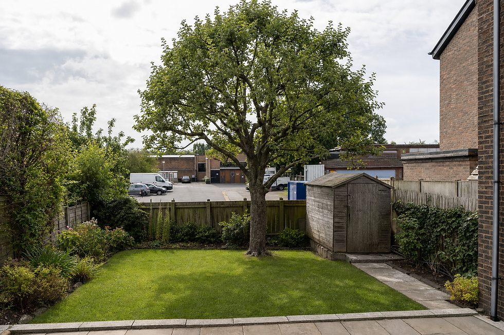 A neatly trimmed garden with a large tree, wooden shed, and a view of parked vehicles in the background.