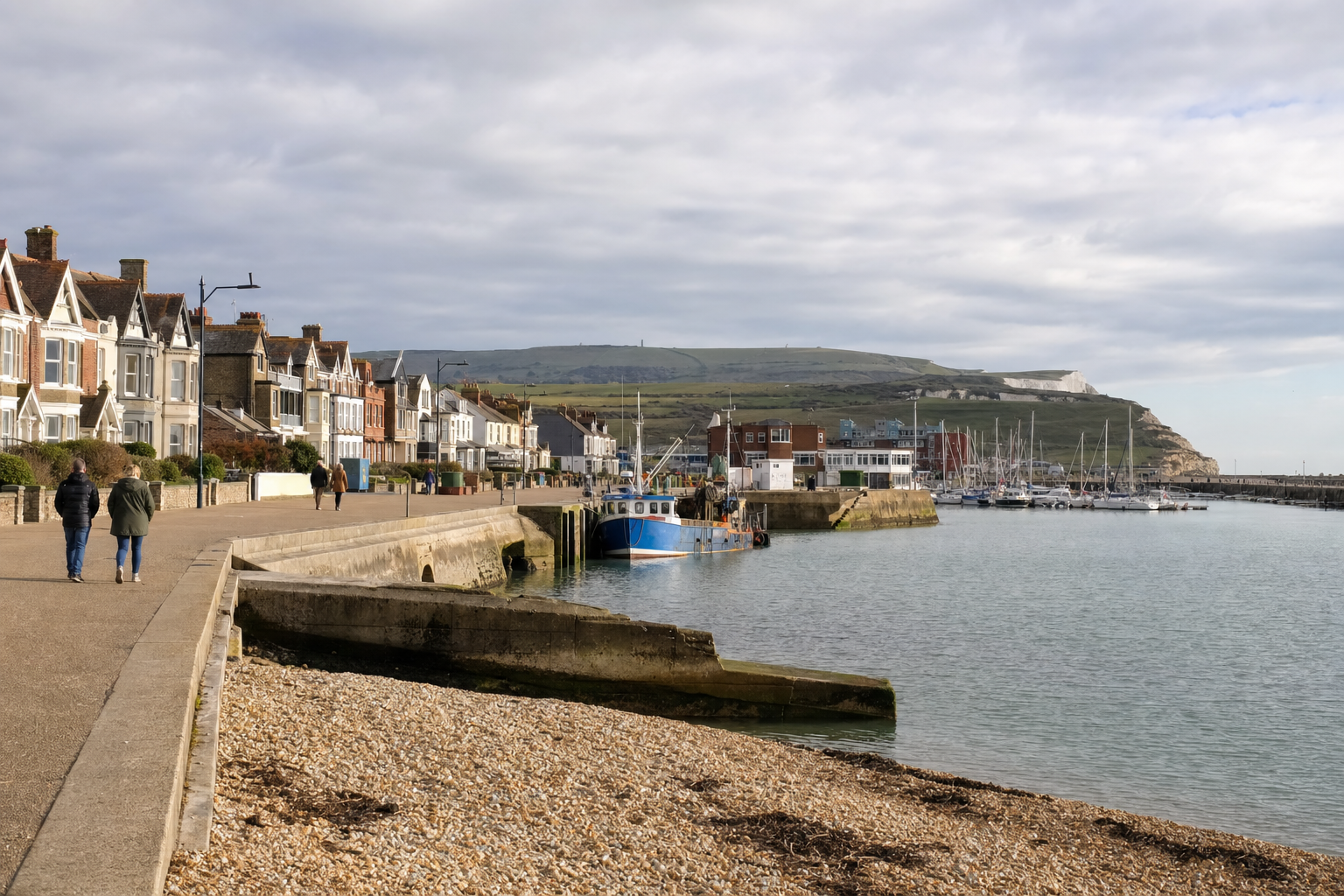 A coastal promenade with people walking, boats docked in the harbour, and houses lining the waterfront under a cloudy sky.