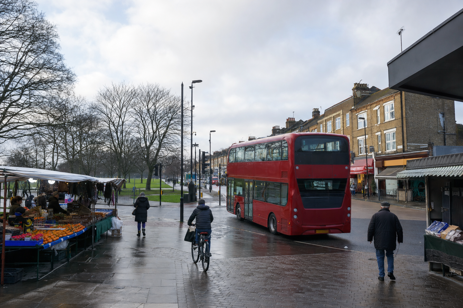 A red double-decker bus passes market stalls on a damp street near a park in a typical British town.