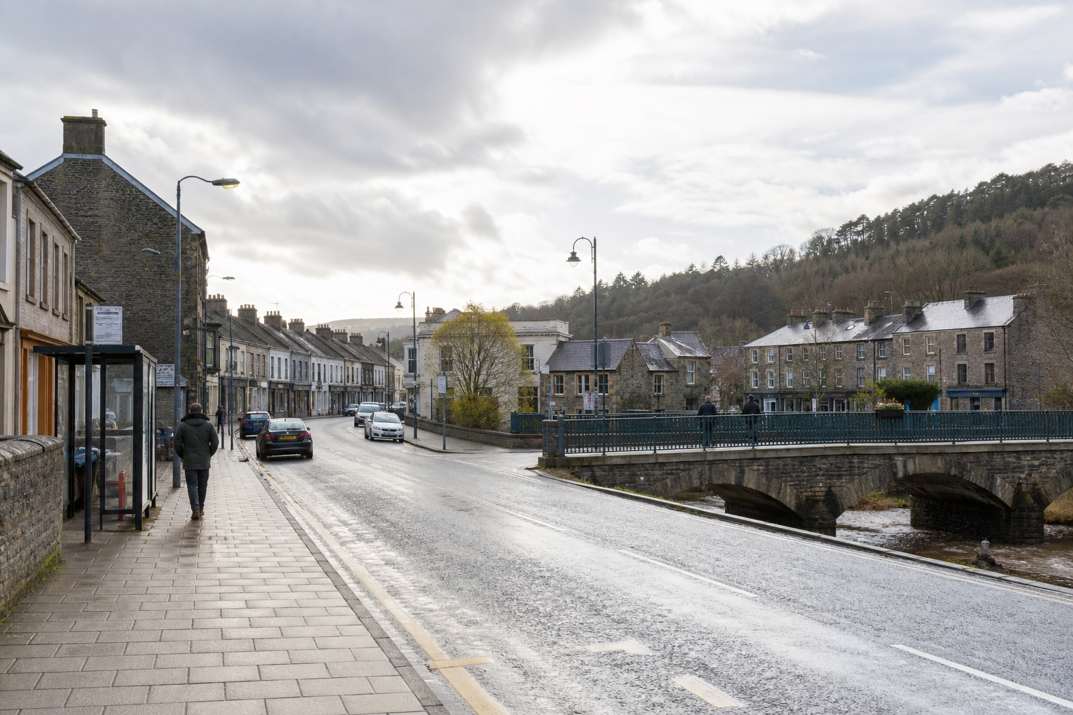 A quiet town street with stone buildings, a bridge over a small river, and overcast skies.