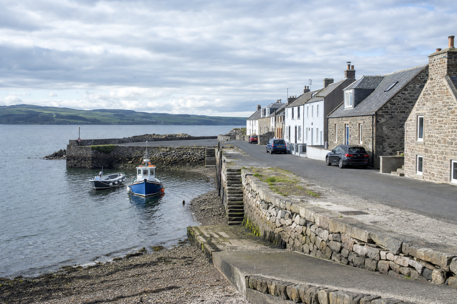 Coastal village street with stone houses, parked cars, and moored boats on a cloudy day.