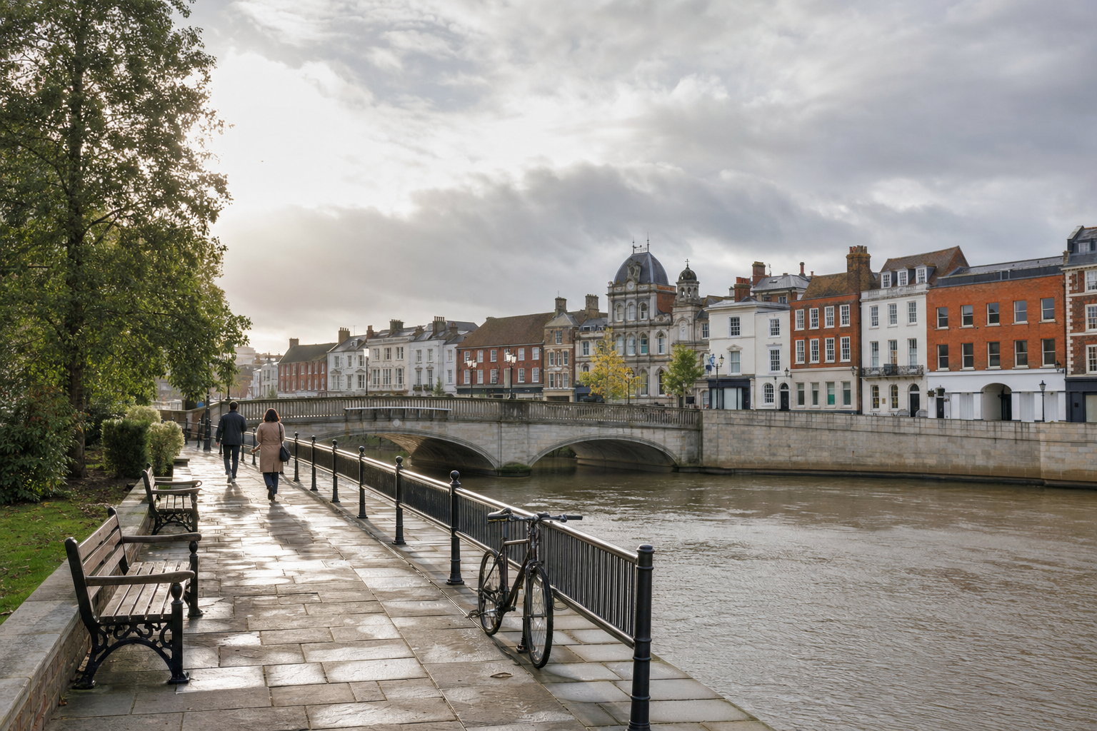 A riverside promenade with benches and a bicycle leads to an elegant bridge in front of historic buildings on a cloudy day.