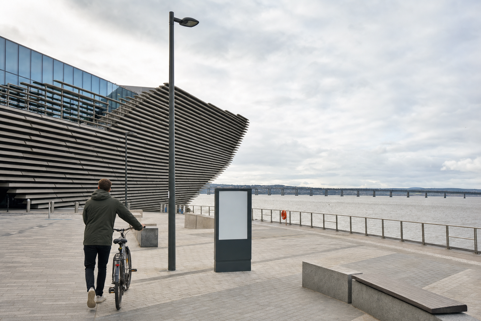 Person walking a bicycle near a modern building by a riverside with a long bridge in the background.