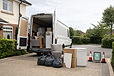 A white moving van parked with its doors open, surrounded by furniture, boxes, and bins on a residential driveway.