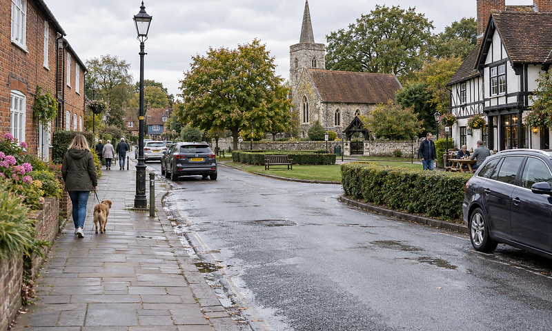 A woman walks a dog along a quaint village street with a church and half-timbered houses nearby.