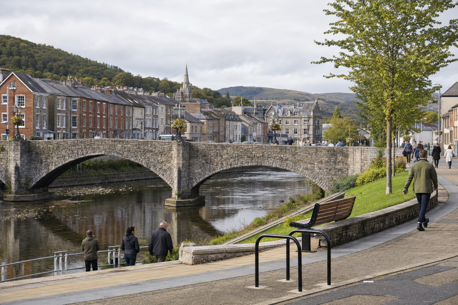 Stone bridge over a river with people walking along a tree-lined path and colourful buildings in the background.