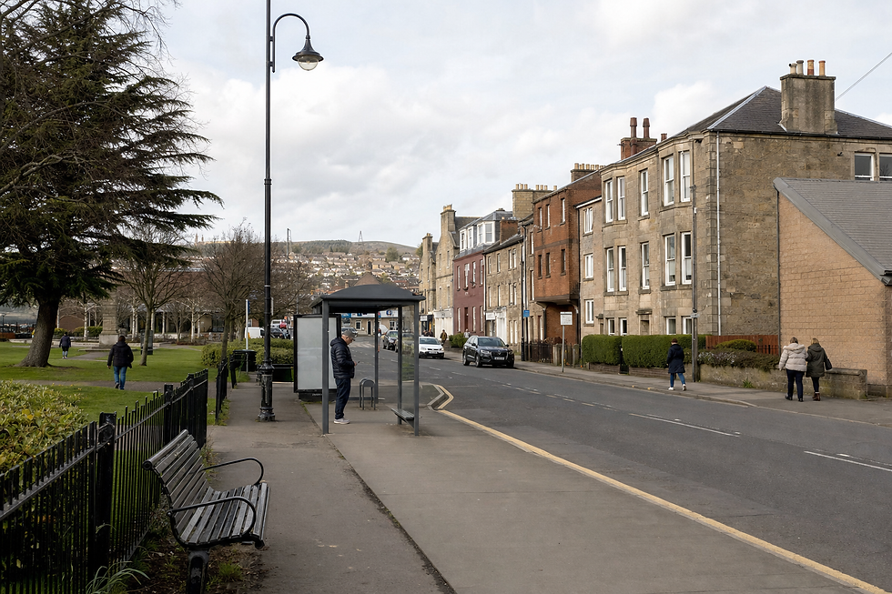A man waits at a bus stop on a quiet street lined with brick buildings and a park on a cloudy day.