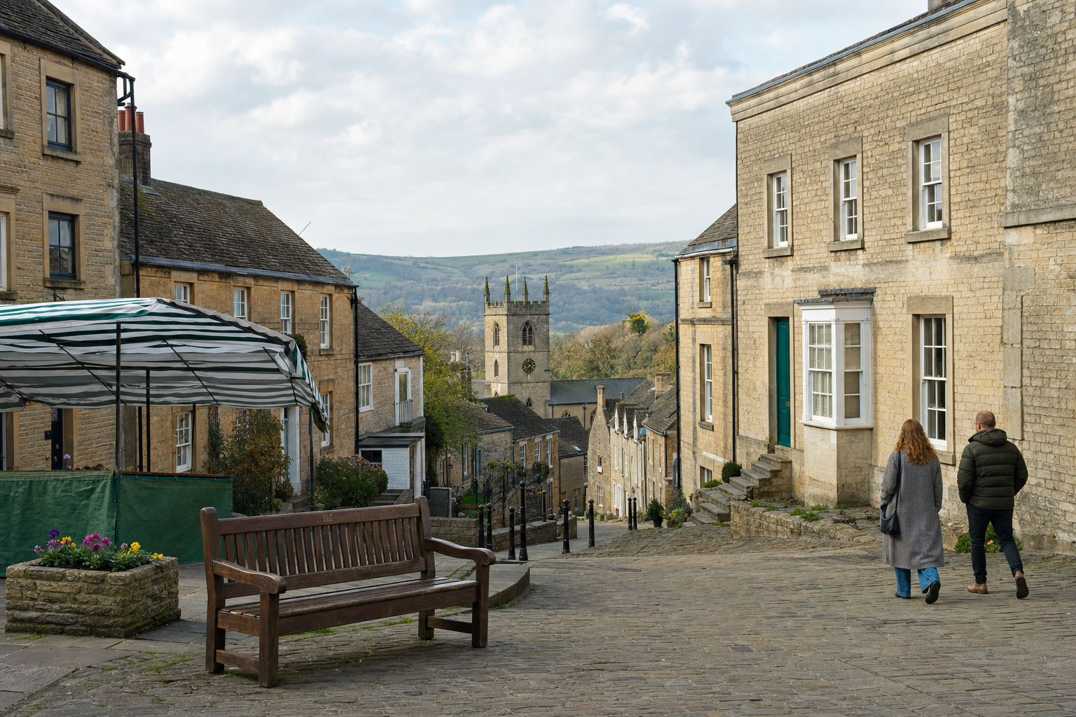 A cobblestone street in a quaint village with old stone buildings and a church tower in the background, two people walking.