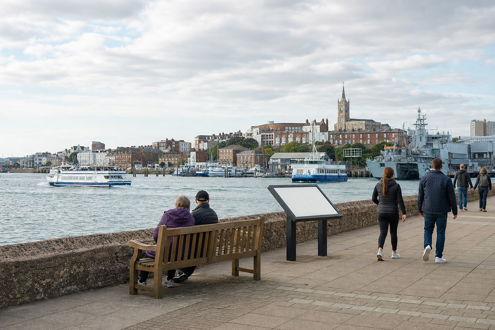 People stroll along a waterfront with benches and a view of ferries and historical buildings in the background.