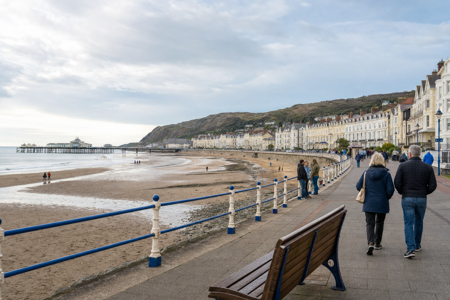 People walking along a seaside promenade with a beach, Victorian buildings, and a pier in the background.