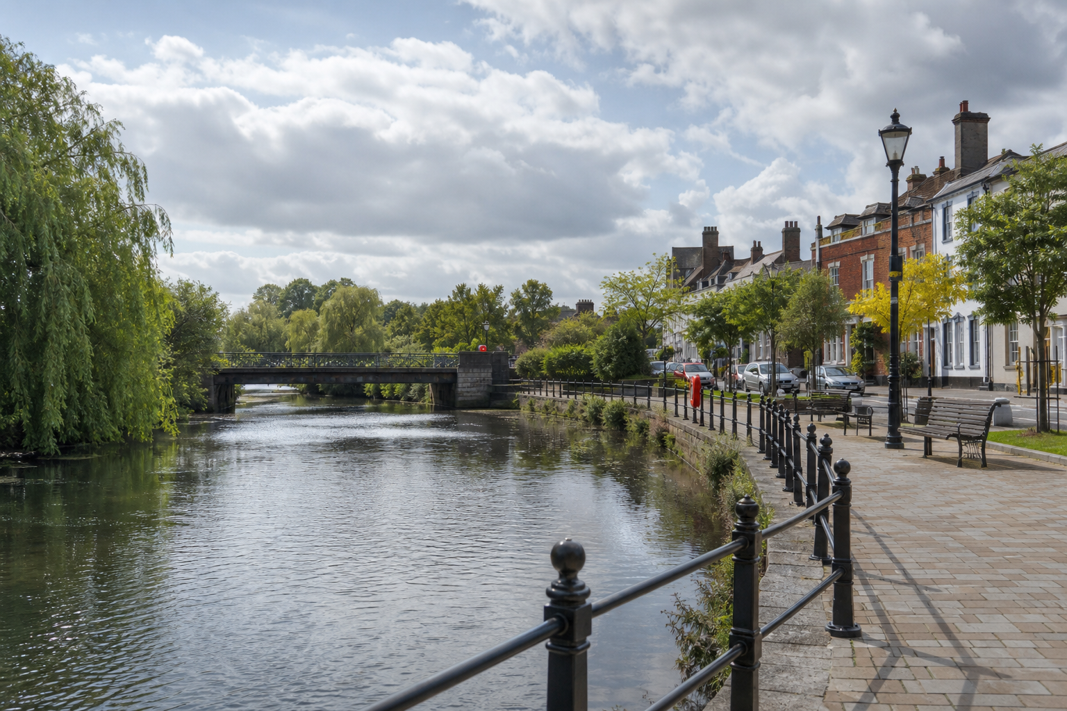 Riverside walkway with benches, trees, and classic houses, leading to a small bridge under a partly cloudy sky.