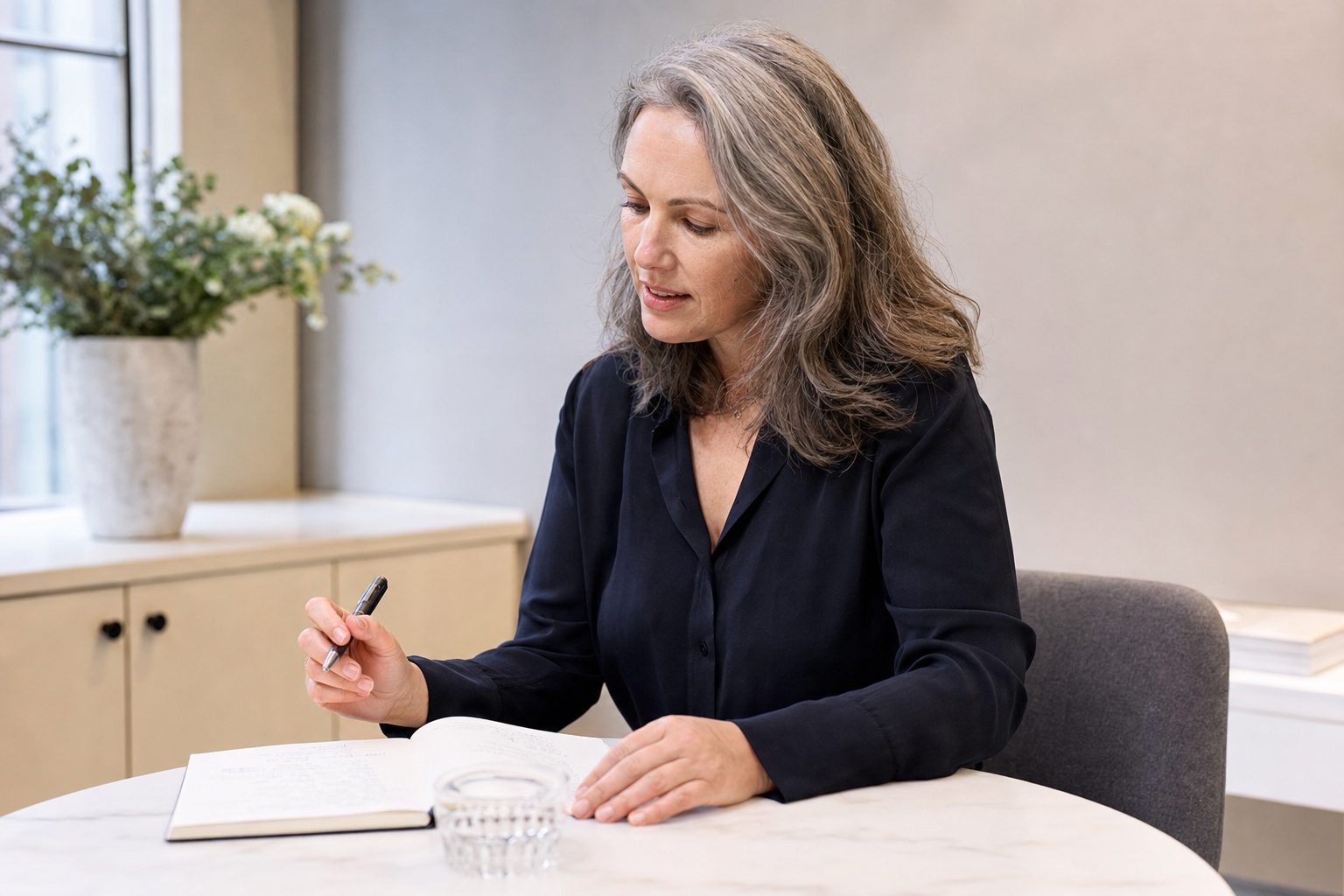 Woman in a dark blouse writing in a notebook at a round table in a bright room.