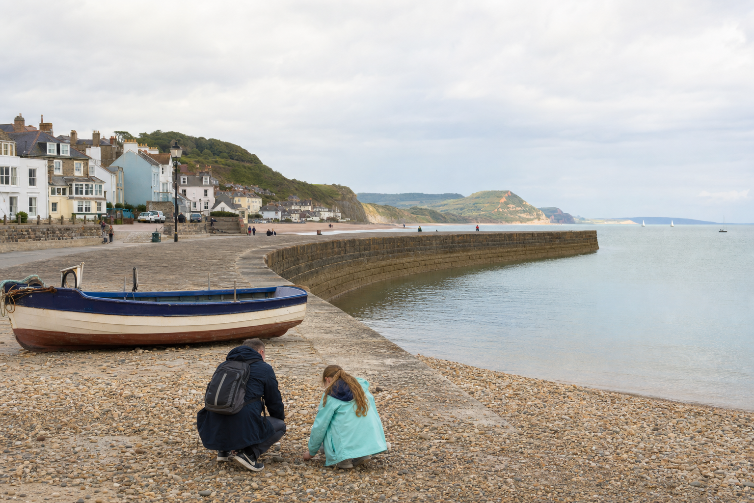 Two people crouching on a pebble beach near a blue boat, with a row of houses and a stone harbour wall in the background.