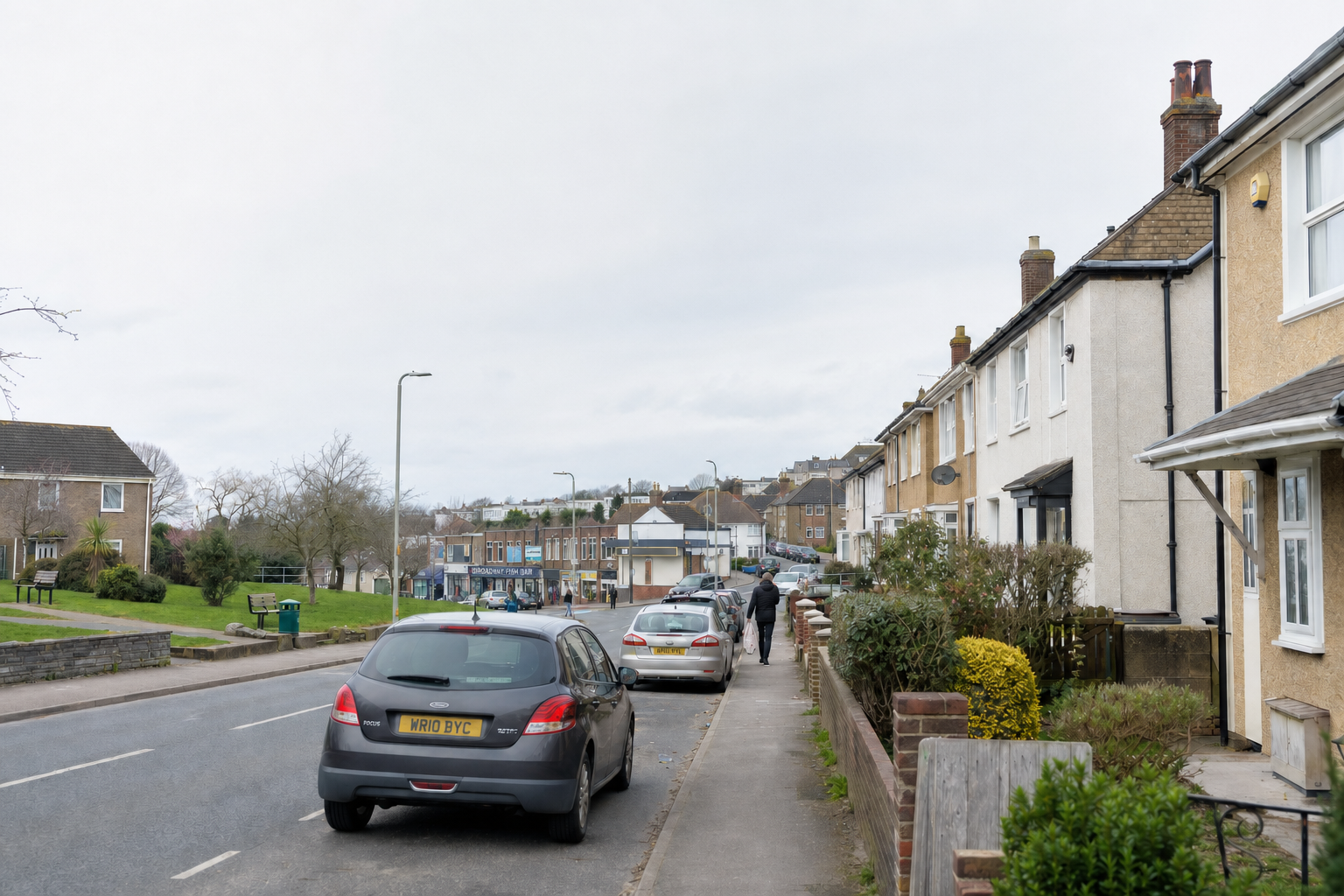Residential street with parked cars and people walking on the pavement, flanked by terraced houses and a small green.