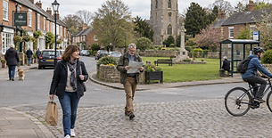 A man and woman walk through a quaint village square, the man reading a map and the woman holding a shopping bag.